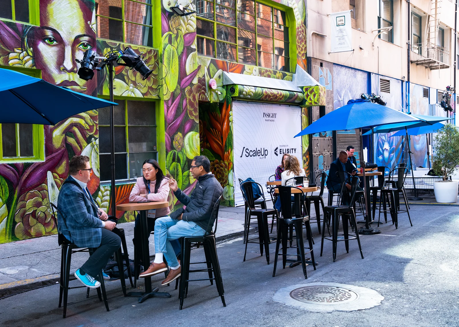 Guests sit at tall outdoor tables beneath blue umbrellas beside a painted mural in Minna Street alley.
